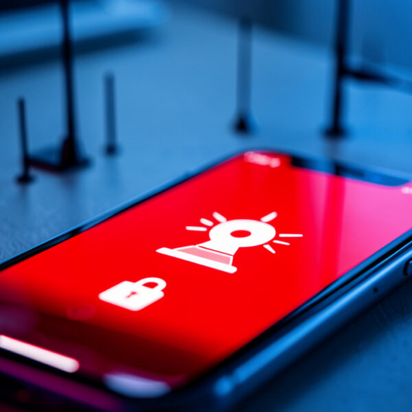 Smartphone verrouillé affichant une interface d’alerte d’urgence rouge avec une icône de sirène, devant des antennes-relais floues.